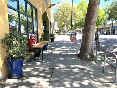 Picture of a sidewalk, in front of the restaurant Gaumenkitzel. To the right the San Pablo Avenue. The sidewalk is shaded by summer trees. In front of the restaurant is a women sitting on a beer garden bench looking towards the photographer.